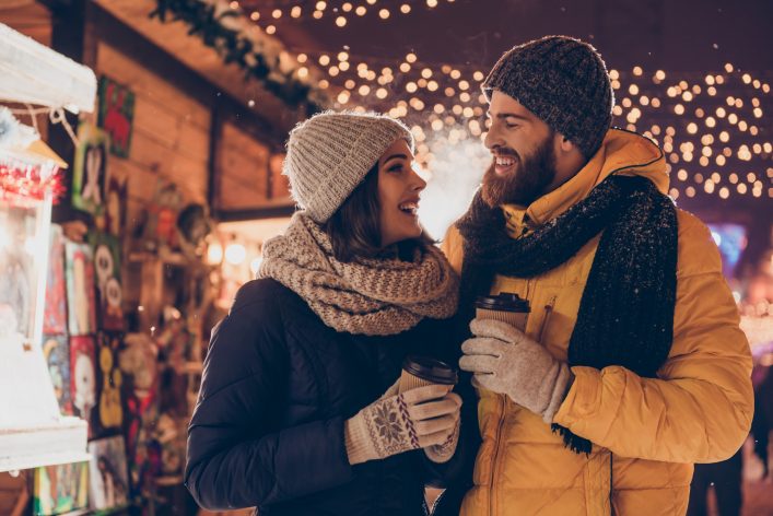 Having-fun-together-at-a-christmas-fairy.-Young-cheerful-couple-is-having-a-walk-with-hot-drinks-enjoying-dressed-warm-looking-at-each-other-and-laugh_shutterstock_714423586